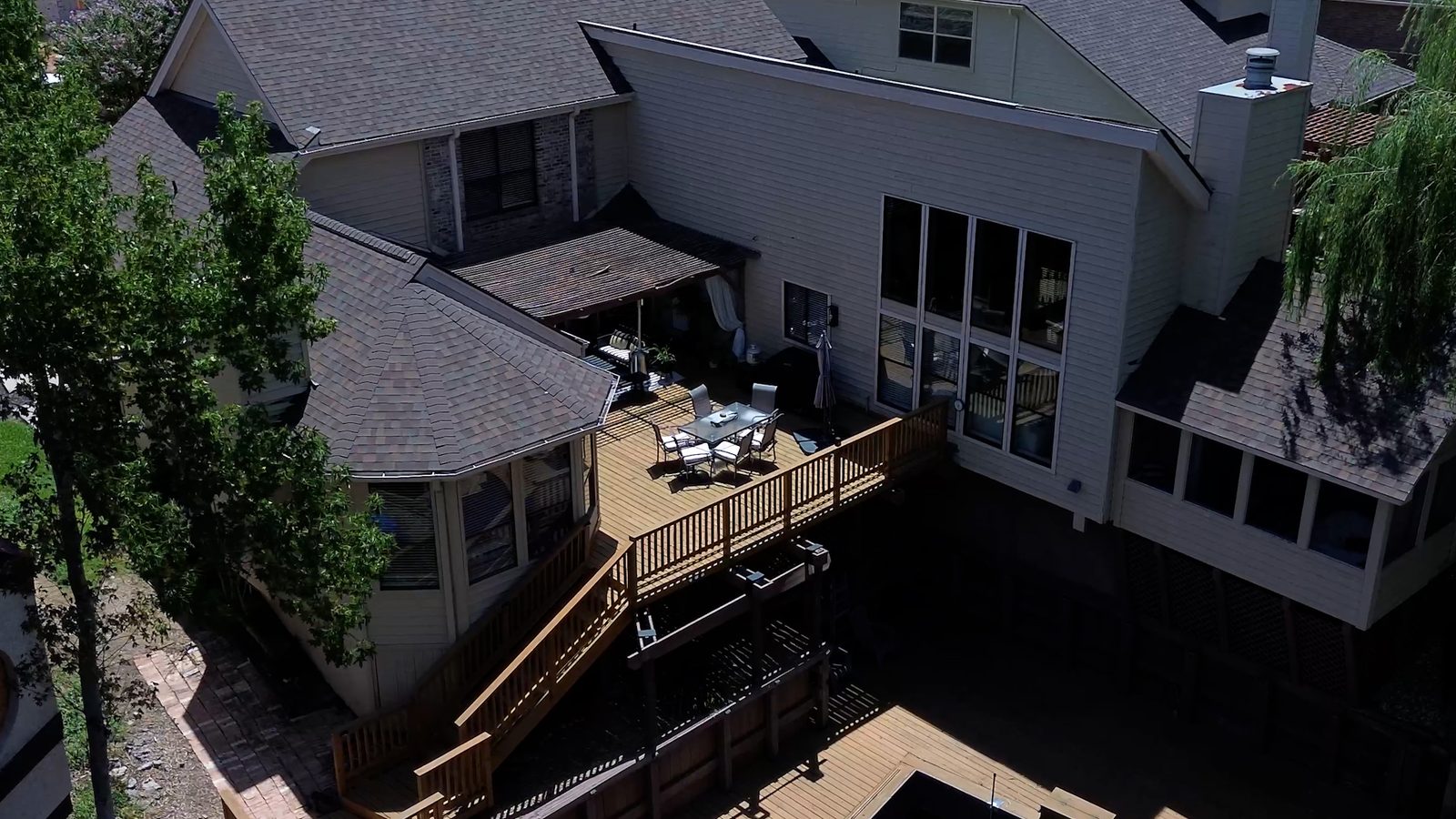 Aerial view of the back of a two-story house, with a raised wooden deck and patio table below a wall of tall windows